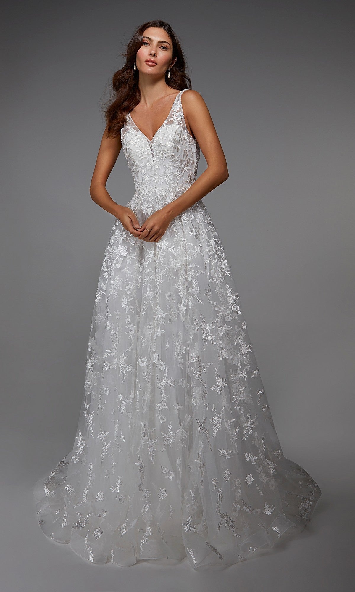 A woman models the Long White Dress: Alyce 7037, a stunning sleeveless gown featuring a V-neckline and intricate floral lace detailing, set against a plain gray background.
